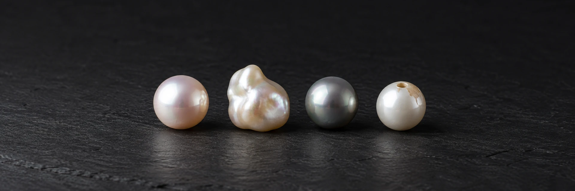 Round white, baroque and gray pearls beside a worn coated bead on dark slate.