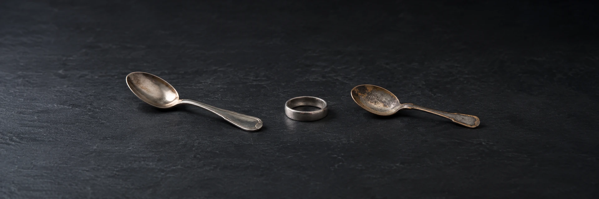 A tarnished silver-colored spoon, plain ring and worn plated spoon on dark slate.